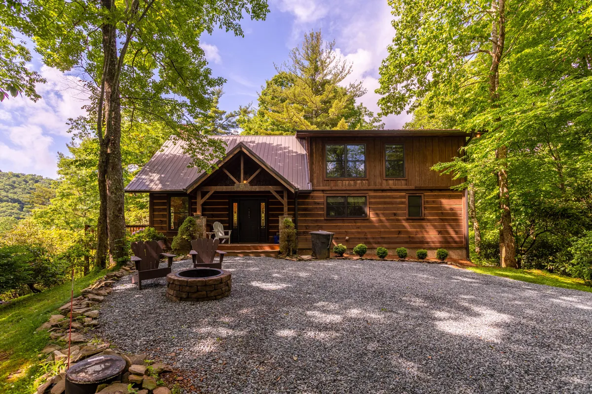 The image features the charming log cabin exterior set amid Boone’s scenic landscape. A gravel driveway leads to the cabin, while a cozy fire pit area is surrounded by tall trees. The setting evokes privacy, tranquility, and outdoor fun, with ample space for gathering, relaxing, and taking in the mountain views.