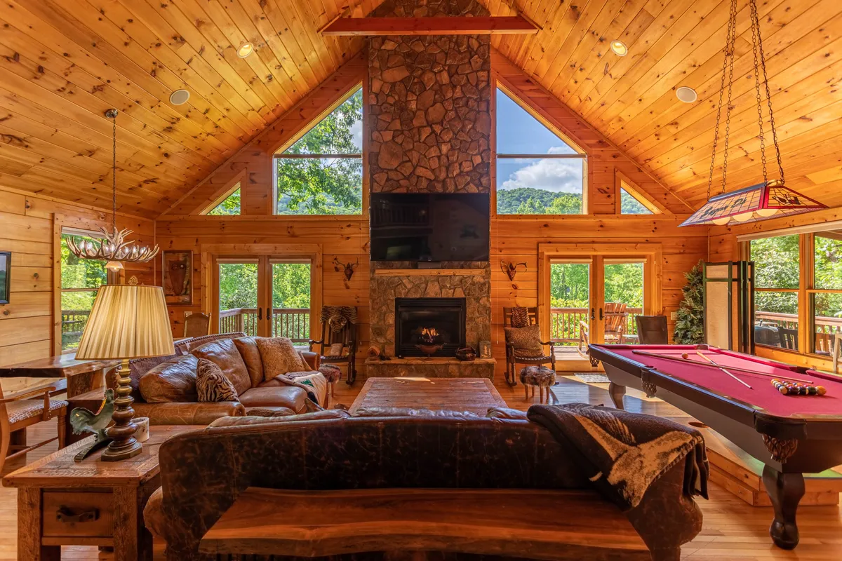 This inviting interior features a large stone fireplace, a pool table, expansive windows framing scenic nature views, and exposed wooden beams. The space is warm and sociable, highlighting areas where guests and their pets can unwind after outdoor adventures or gather for cozy evenings together.