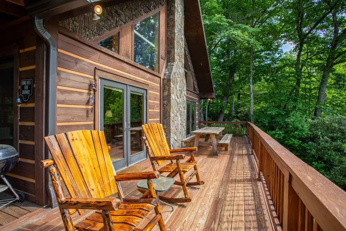A tranquil wooden deck area featuring Adirondack-style wooden chairs, a sturdy stone wall, and large windows that reflect the natural beauty outside. The space is set up for relaxation, socializing, and enjoying the sights and sounds of the surrounding wilderness.