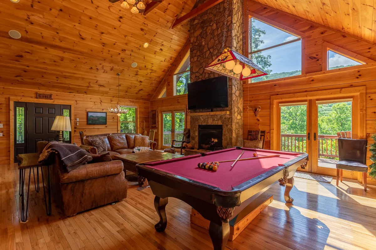 This image centers on the game area within the great room, with a pool table in the foreground, a stone fireplace, large windows letting in natural light, and a comfortable seating area. The setting is ideal for game nights, socializing, and appreciating the views, capturing the playful spirit described in the section.