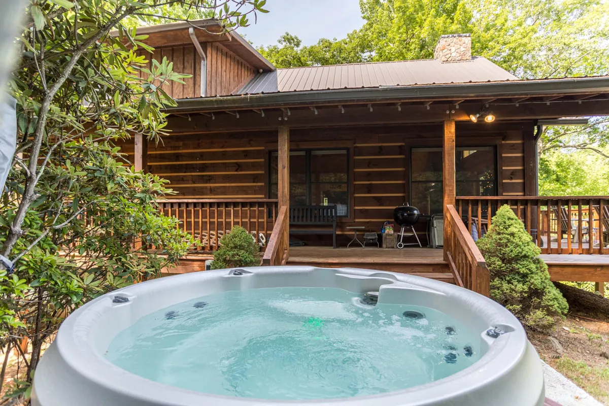 A hot tub sits prominently on a wooden porch, framed by lush greenery and the rustic log cabin exterior. The tranquil scene suggests cozy evenings spent soaking while surrounded by mountain beauty.