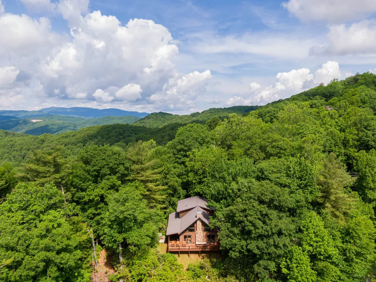 A view of the wooden cabin nestled among tall trees with a dramatic mountain backdrop and clouds drifting by. The scene conveys seclusion and easy access to nature, ideal for families looking to explore the great outdoors. The surrounding forest hints at nearby trails and adventures, while the inviting porch encourages relaxation after a day of activity.