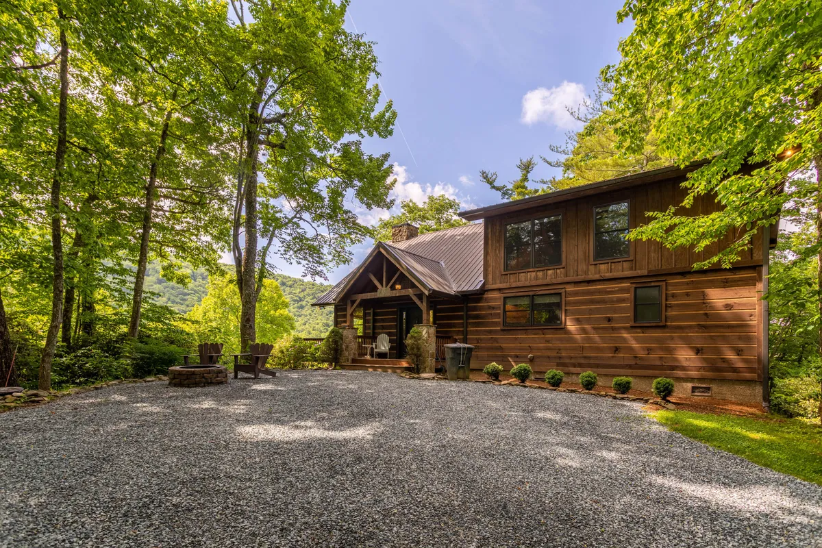 A wooden cabin is nestled among tall trees, featuring an outdoor seating area and a gravel driveway. There’s a clear view of the fire pit area, where guests can gather for storytelling under the stars, and plenty of green space for pets to explore.