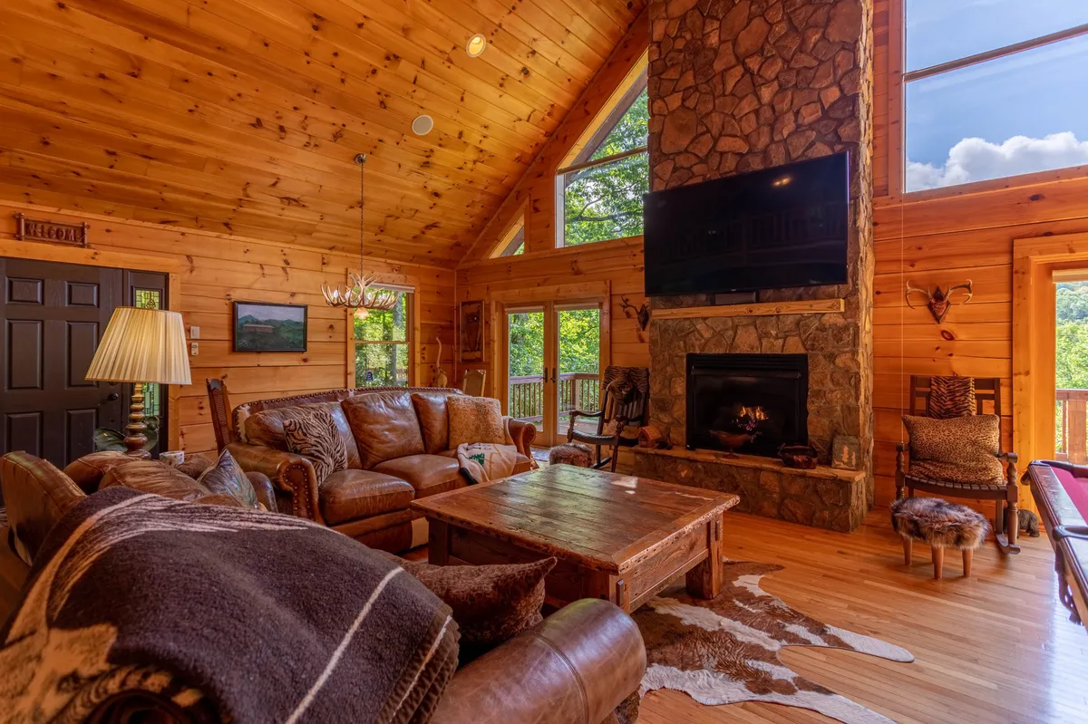 A spacious family area centers around a stone fireplace, complemented by large windows offering sweeping views of the surrounding mountains. The inviting wooden furniture and chandelier add a rustic charm, emphasizing memorable moments with loved ones and pets.