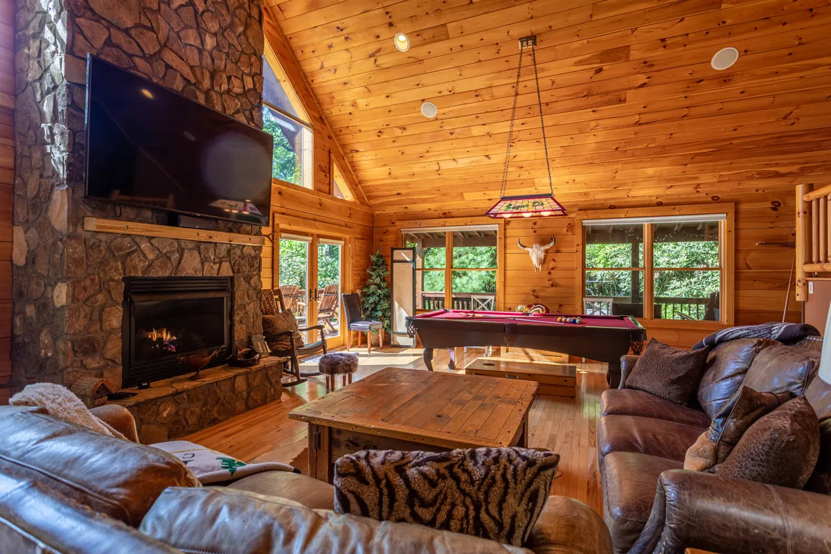 A cozy, light-filled room with a stone fireplace, pool table, and panoramic windows looking out onto natural vistas. The wooden beams and relaxed decor foster a welcoming retreat suitable for both humans and pets.