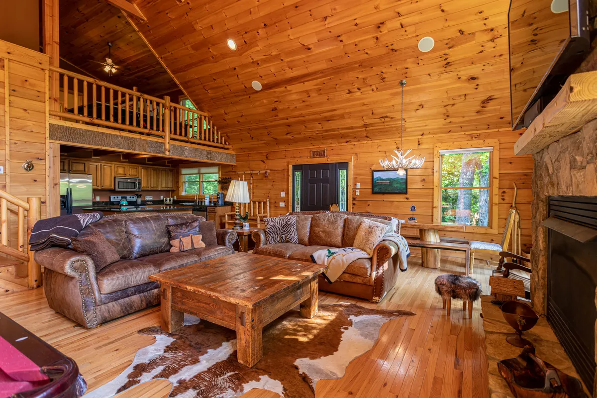 A cozy angle of the great room featuring plush leather sofas positioned around a fireplace, a large window offering views of the surrounding landscape, and a visible kitchen area in the background. The space radiates warmth and togetherness, inviting guests to relax or share stories.