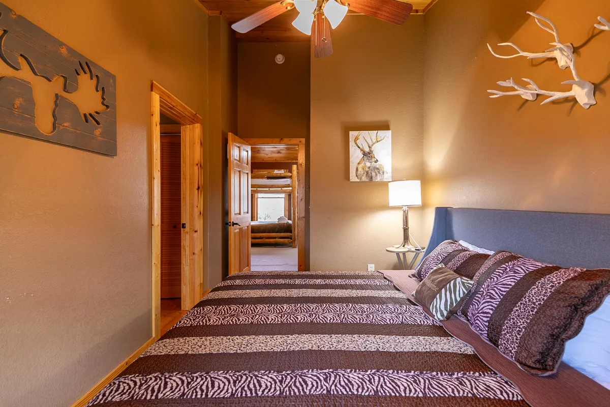 A rustic bedroom with a king-sized bed adorned with patterned bedding, complemented by wooden wall art, a bedside lamp, and a painted animal portrait. Gentle natural light filters in, creating an inviting, tranquil retreat ideal for restful sleep or quiet reading.