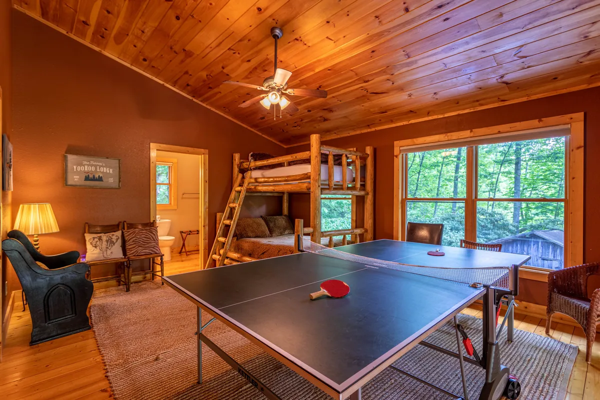 A vibrant loft space featuring a ping pong table at the center, flanked by a cozy bunk bed and large windows that invite natural light. Young guests can be seen playing ping pong, while others relax on a comfortable couch or read by the window, with playful decor and a relaxed atmosphere throughout.