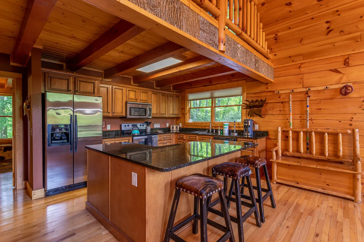 A cozy, welcoming kitchen featuring a large island with granite countertops, stainless steel appliances, rich wooden cabinetry, and bar stools. Ample natural light streams in through generous windows, highlighting the practical and stylish design. The open layout invites gathering and conversation while cooking.